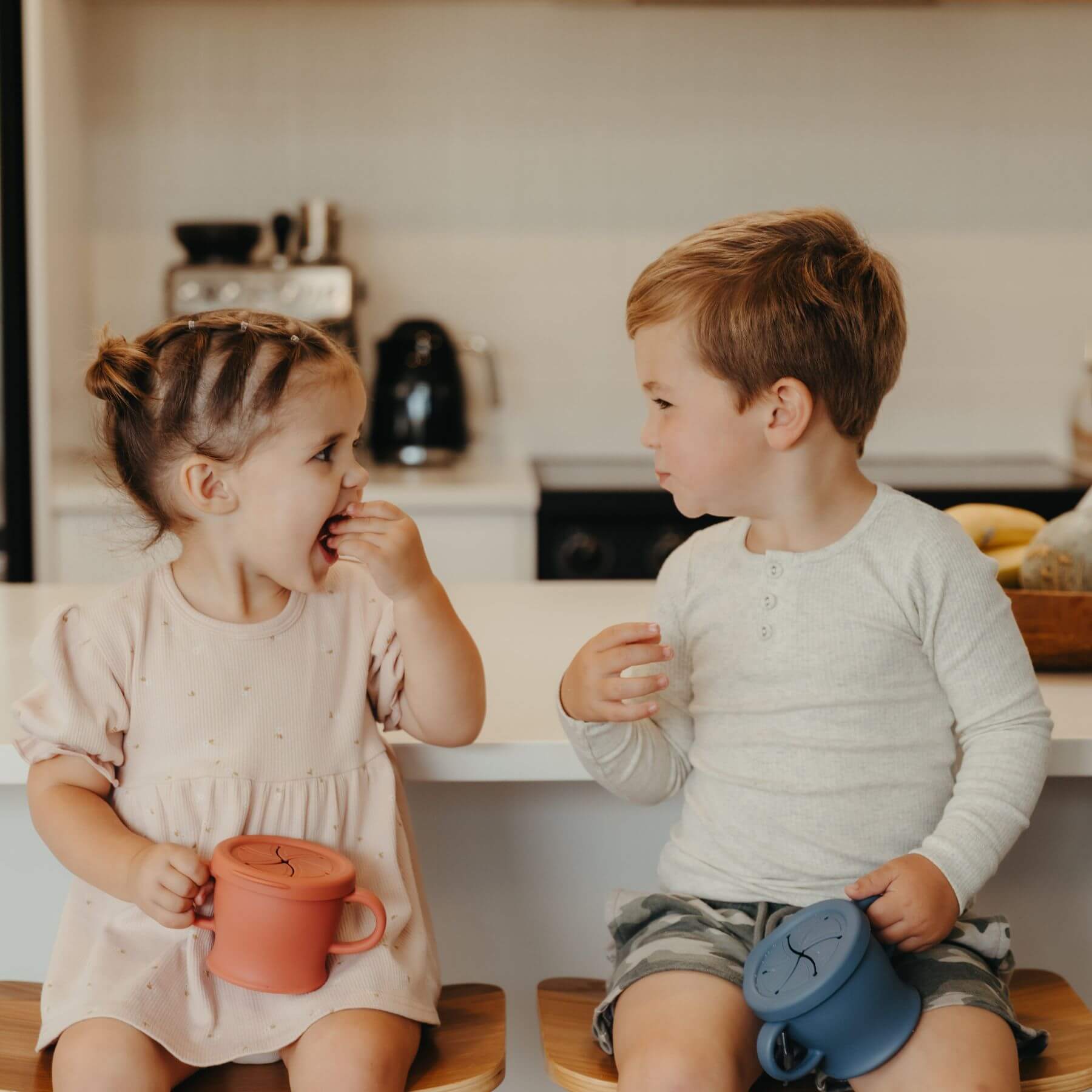 Two children sitting on a kitchen counter, holding Haakaa Silicone Sip-N-Snack Cup 8 oz