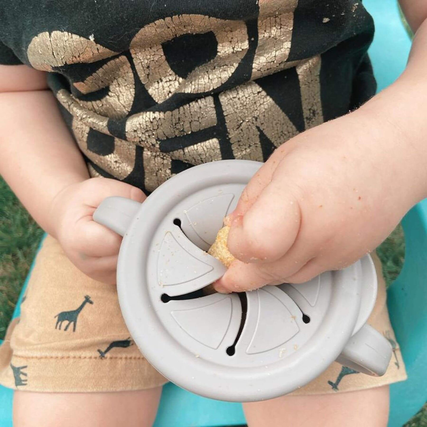 Child holding Haakaa Silicone Sip-N-Snack Cup 8 oz, wearing a black shirt with gold text.