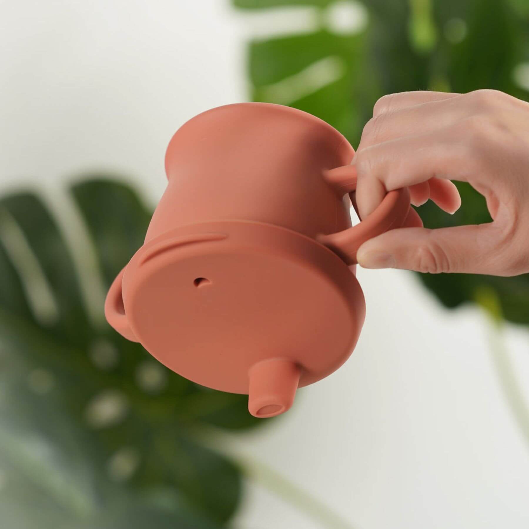Haakaa Silicone Sip-N-Snack Cup 8 oz held by a hand with a blurred green plant background