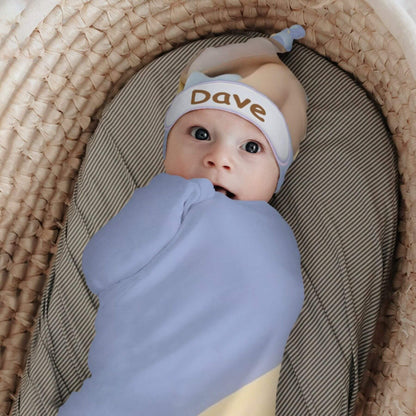 A baby swaddled in a light blue blanket and wearing a matching light blue hat with the name 'Dave' printed in brown, lying in a woven bassinet on a striped mattress.