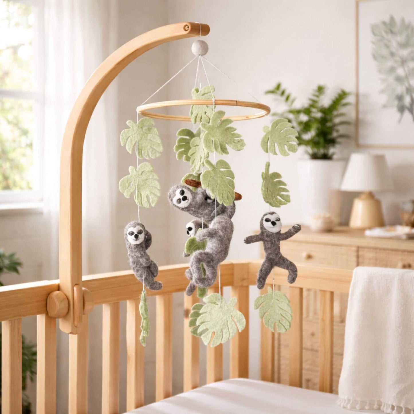 Baby crib with Ganapati Crafts Co. Sloth Baby Mobile hanging above, in a room with plants and a lamp.