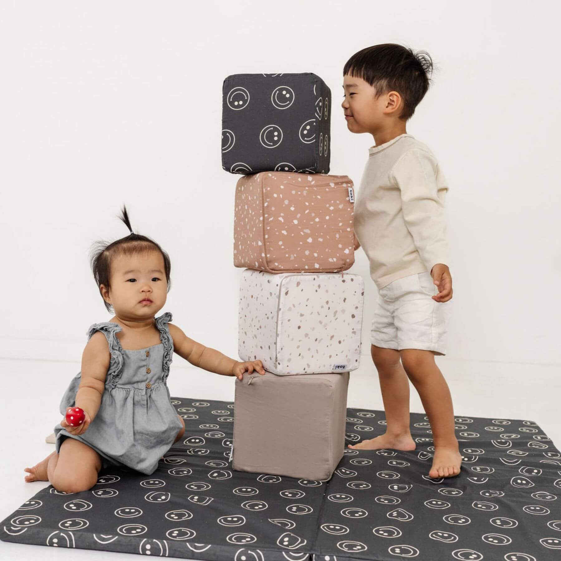 Two children playing with a stack of patterned cushions on a white background