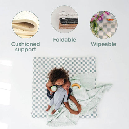 Checkered crib sheet with child on a white background, featuring 'Cushioned support', 'Foldable', and 'Wipeable' text.