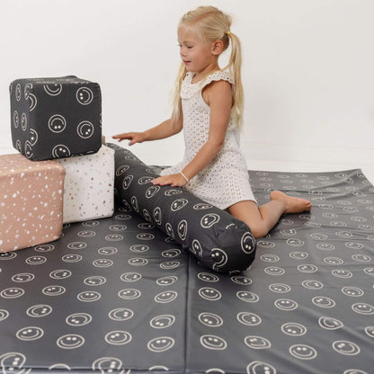 Child playing on a gray mat with smiley face patterns, surrounded by colorful blocks.