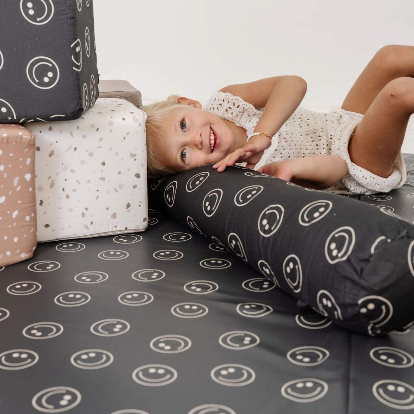 Child lying on a bed with smiley face pattern, surrounded by pillows with similar designs.