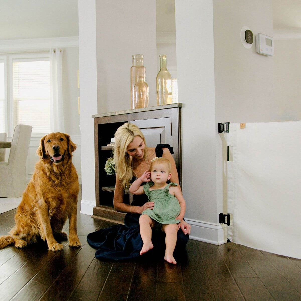 A woman sits on a dark wood floor in a bright home interior, holding a baby dressed in a green outfit while a golden retriever dog sits to her left. A white baby gate is visible on the right side of the room.