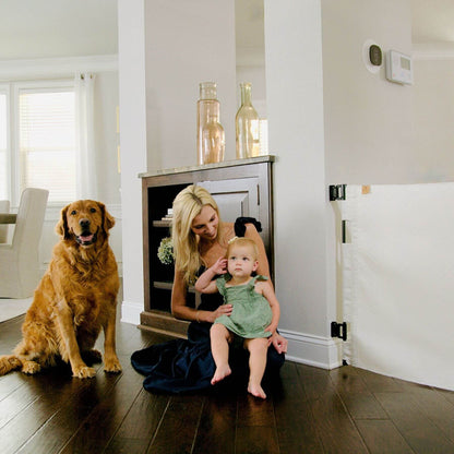 A woman sits on a dark wood floor in a bright home interior, holding a baby dressed in a green outfit while a golden retriever dog sits to her left. A white baby gate is visible on the right side of the room.