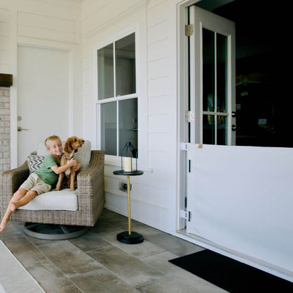 A child sits in a wicker armchair on a white porch, hugging a small dog. A white door with a white bottom gate is visible to the right, along with a small side table and gray stone tile flooring.