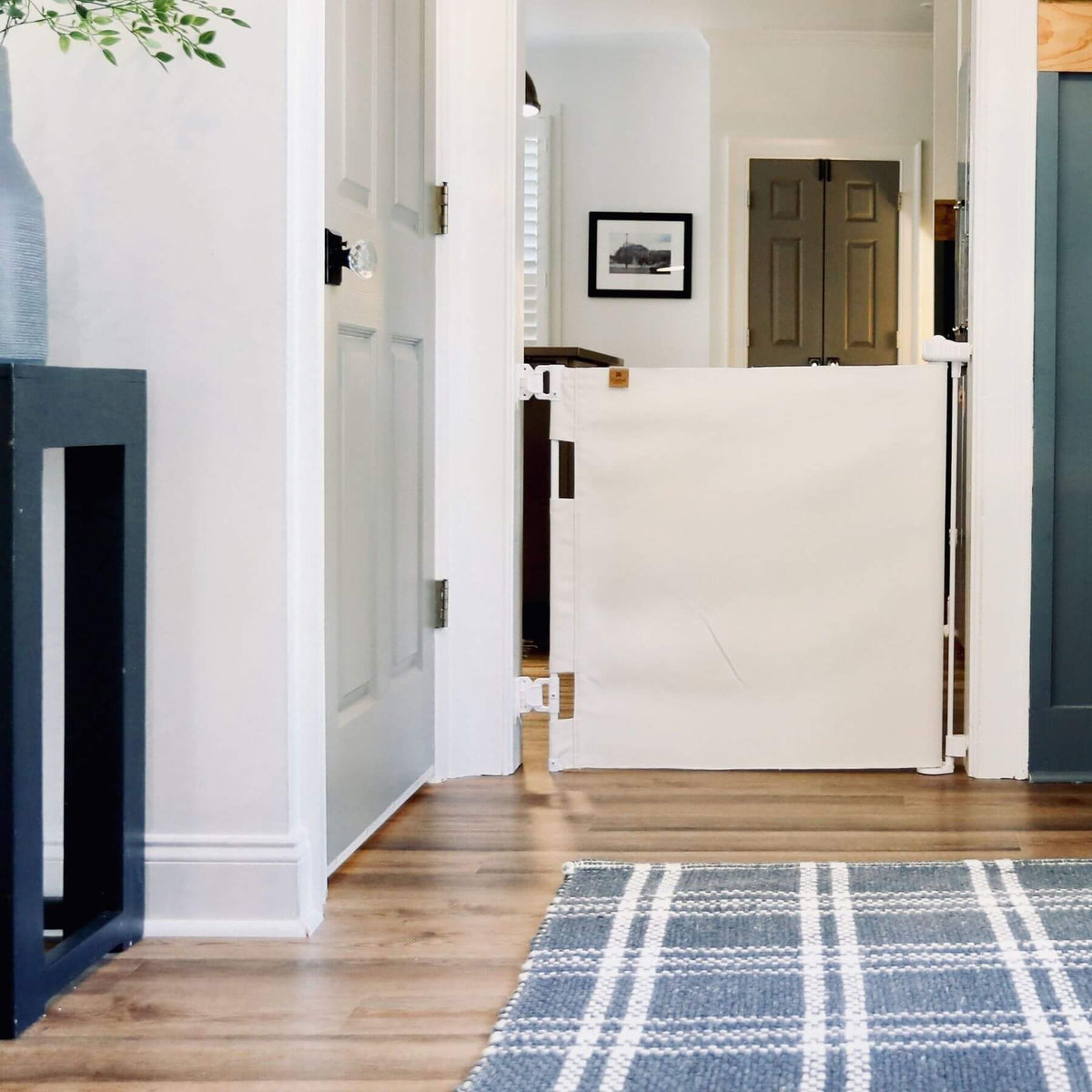 A white fabric baby safety gate installed on a wooden floor in a home hallway, with a blue and white plaid rug in the foreground.