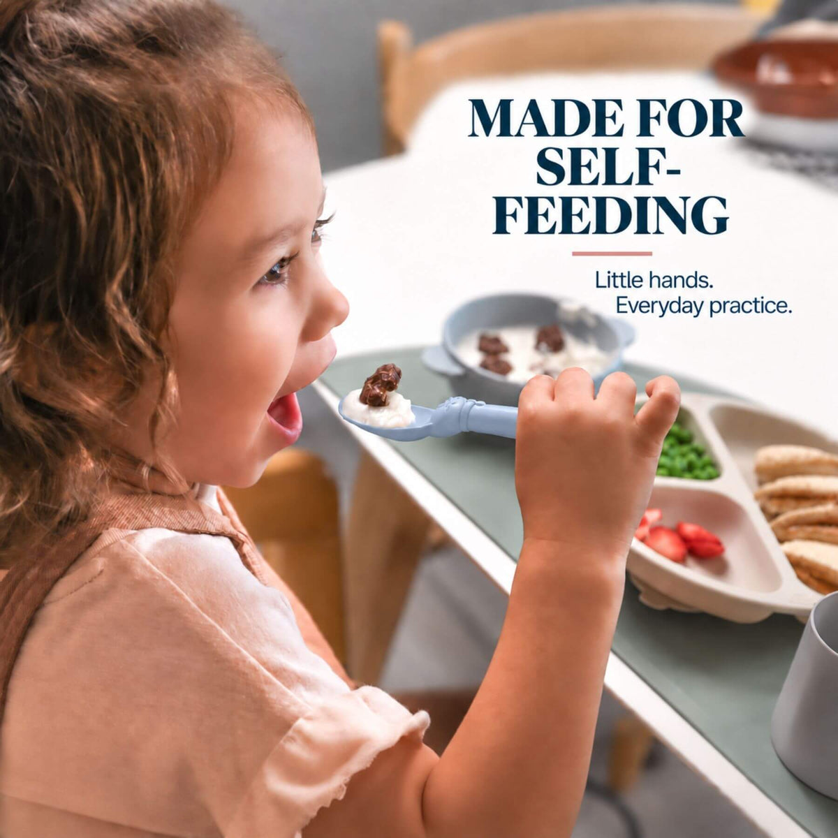 A young child eating from a light blue self-feeding spoon at a table, with the text 'MADE FOR SELF-FEEDING' and 'Little hands. Everyday practice.