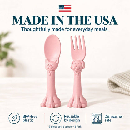 Spoon la la Toddler Utensils — 2-Pack - Pink, shown on a light tabletop with a softly blurred kitchen background. Large text at the top reads 'MADE IN THE USA' with an American flag, and feature icons at the bottom mention BPA-free plastic, reusable by design, and dishwasher safe.