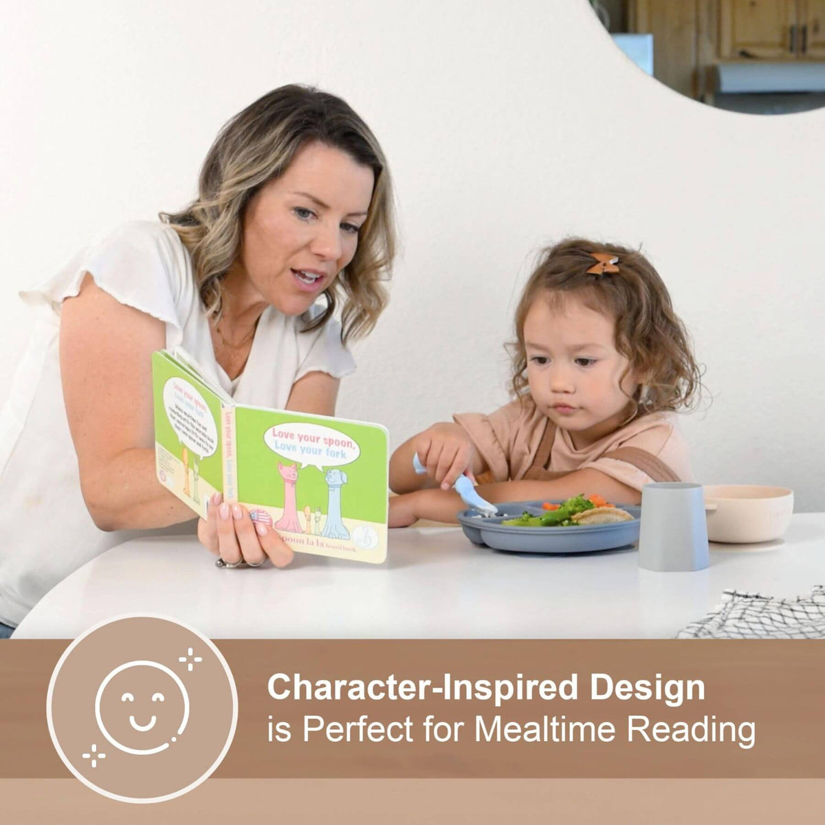 A woman and a young child sit at a table reading a colorful children's book. The woman holds an open illustrated page while the child sits beside her with a plate of food and a spoon.