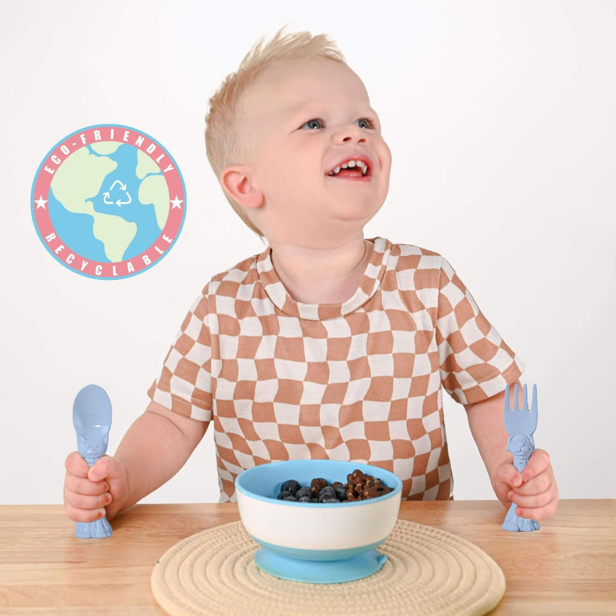 A smiling toddler sits at a table holding a blue plastic spoon and fork, with a blue-and-white bowl of blueberries in front.