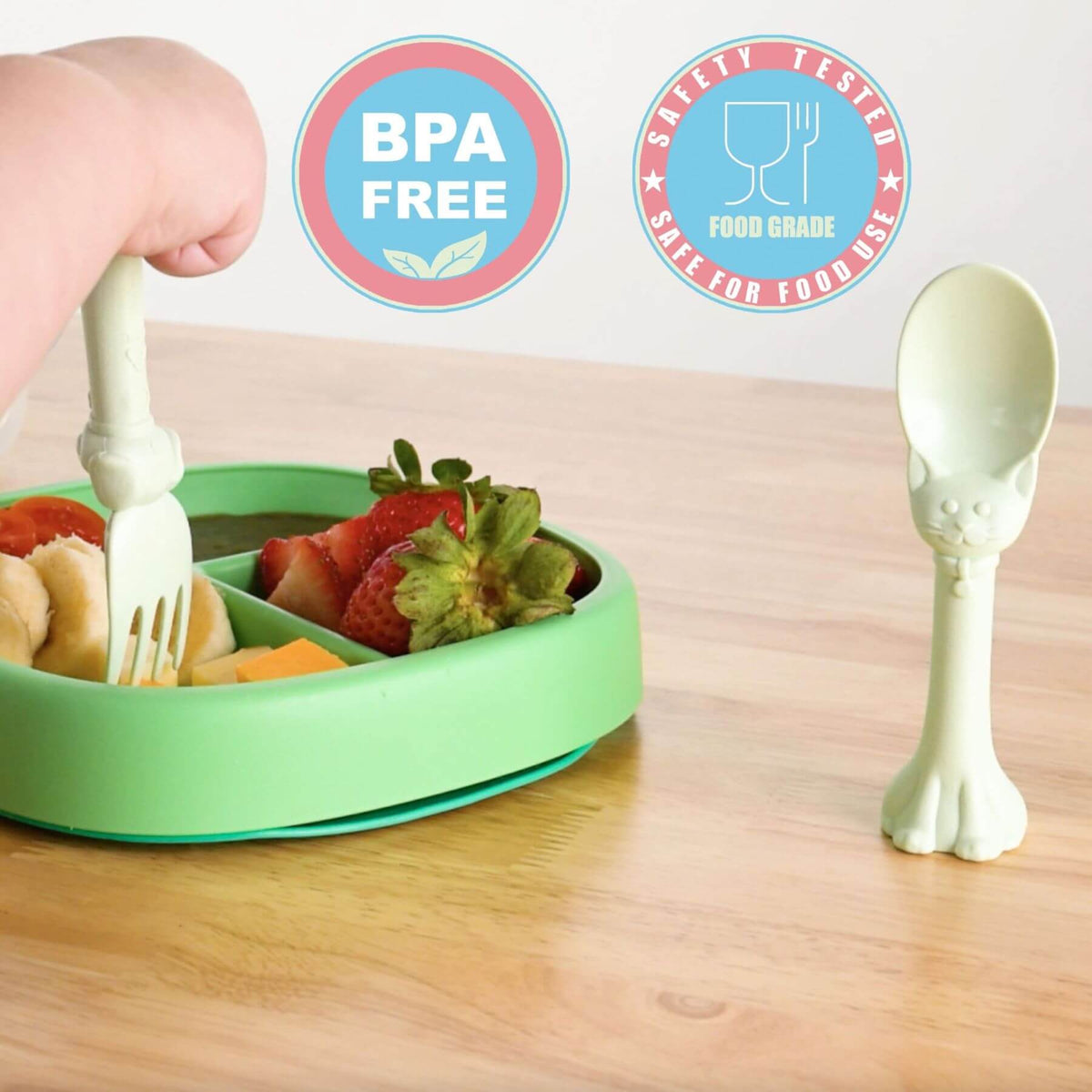 A light green baby mealtime plate with compartments, shown with a hand using a matching bone-shaped utensil to serve fruit. A second bone-shaped spoon is shown standing on the wooden table.