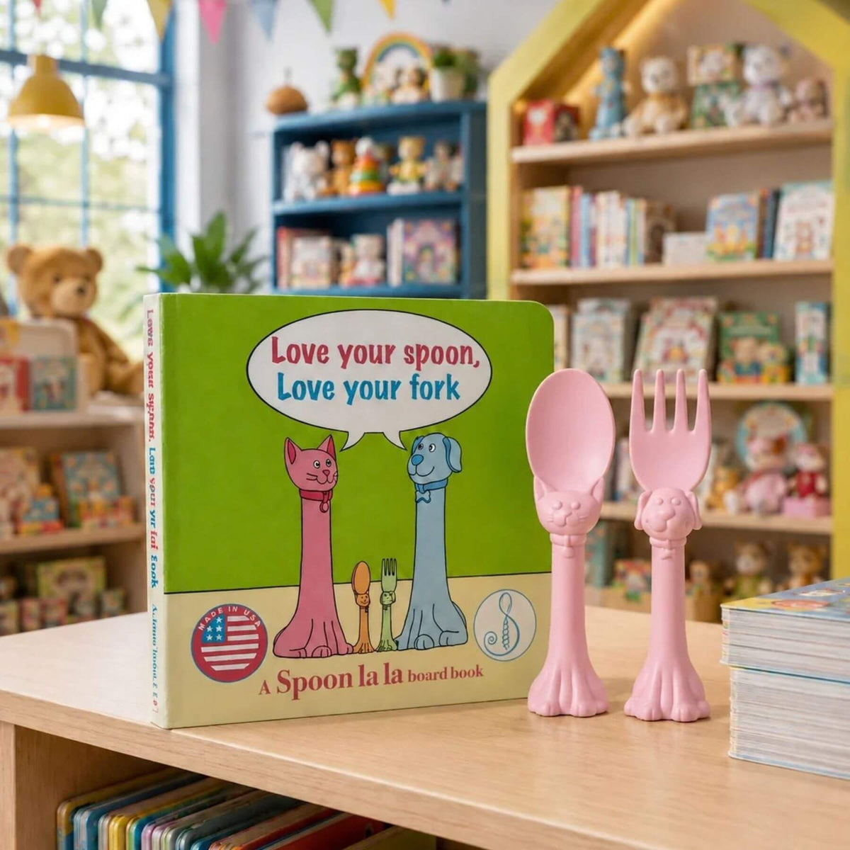 A children's board book titled 'Love your spoon, Love your fork' stands on a wooden table beside a pink spoon and matching pink spork, with a softly blurred nursery or children's room background.