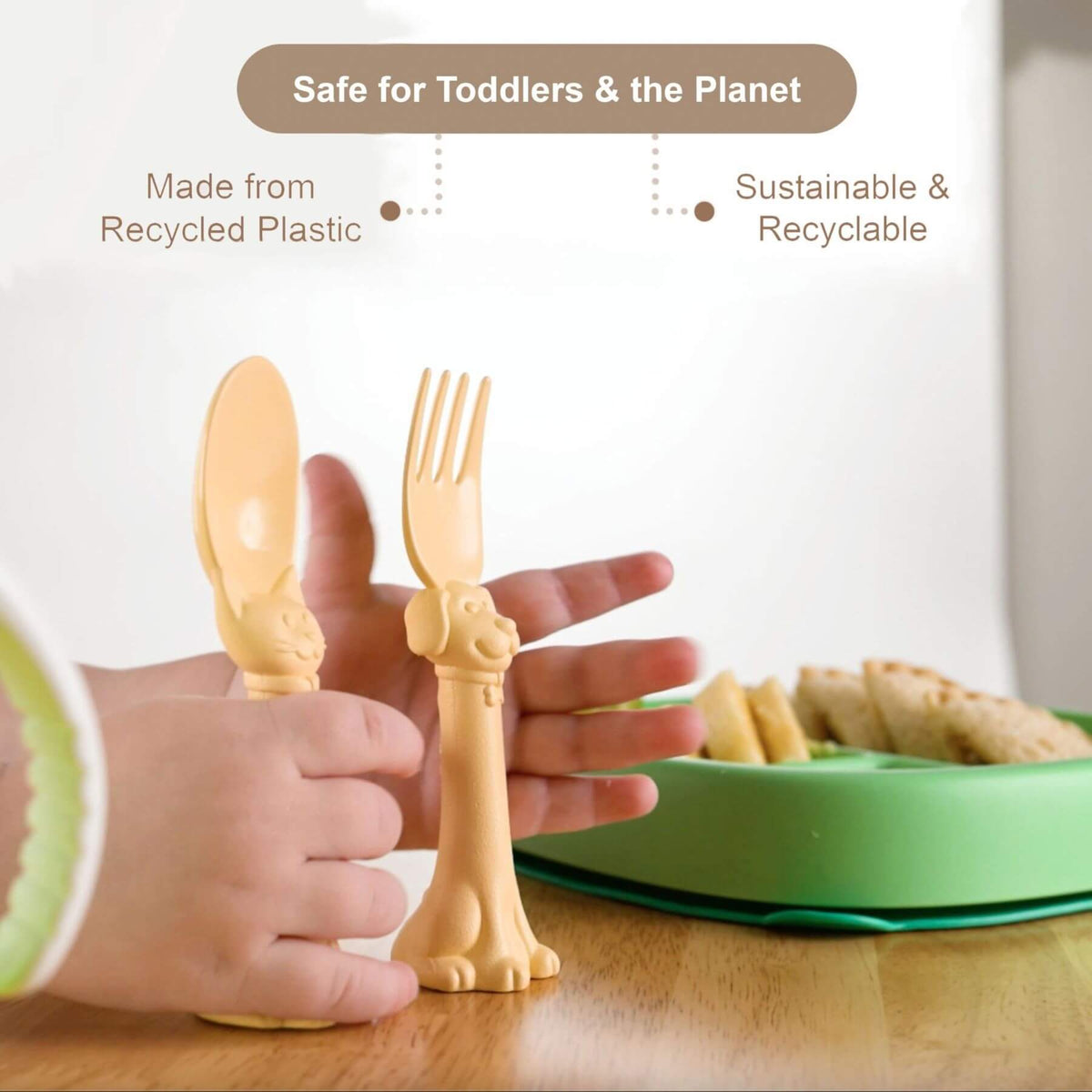 Toddler-friendly plastic spoon and fork set shaped like animal heads, held by a child's hands on a wooden table with a green plate in the background. Text overlays highlight recycled plastic and sustainability.
