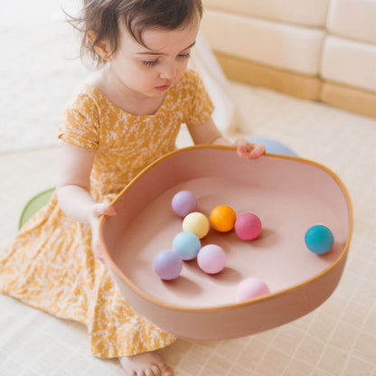 Child playing with colorful balls in a pink bowl on a light-colored floor.