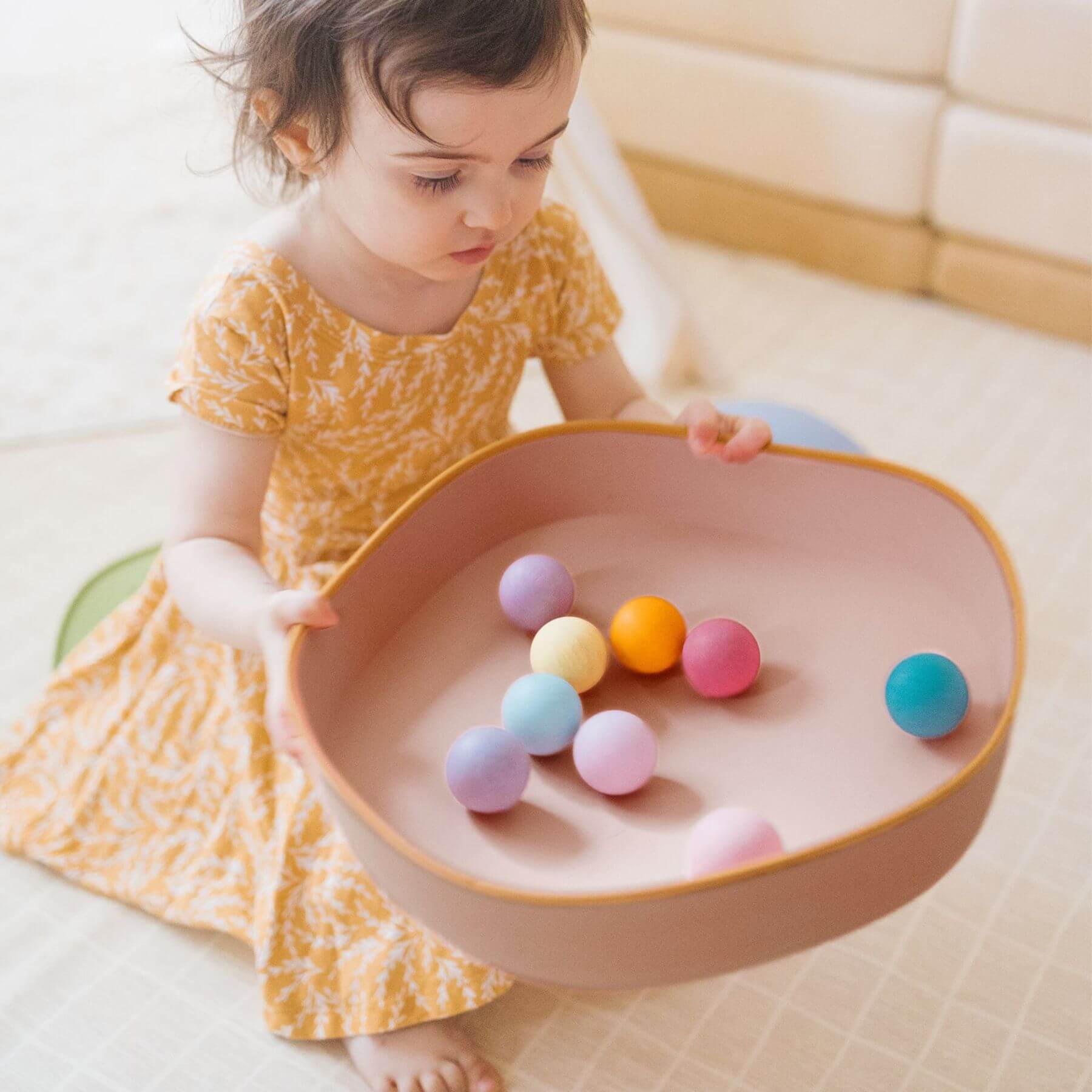 Child playing with colorful balls in a pink bowl on a light-colored floor.