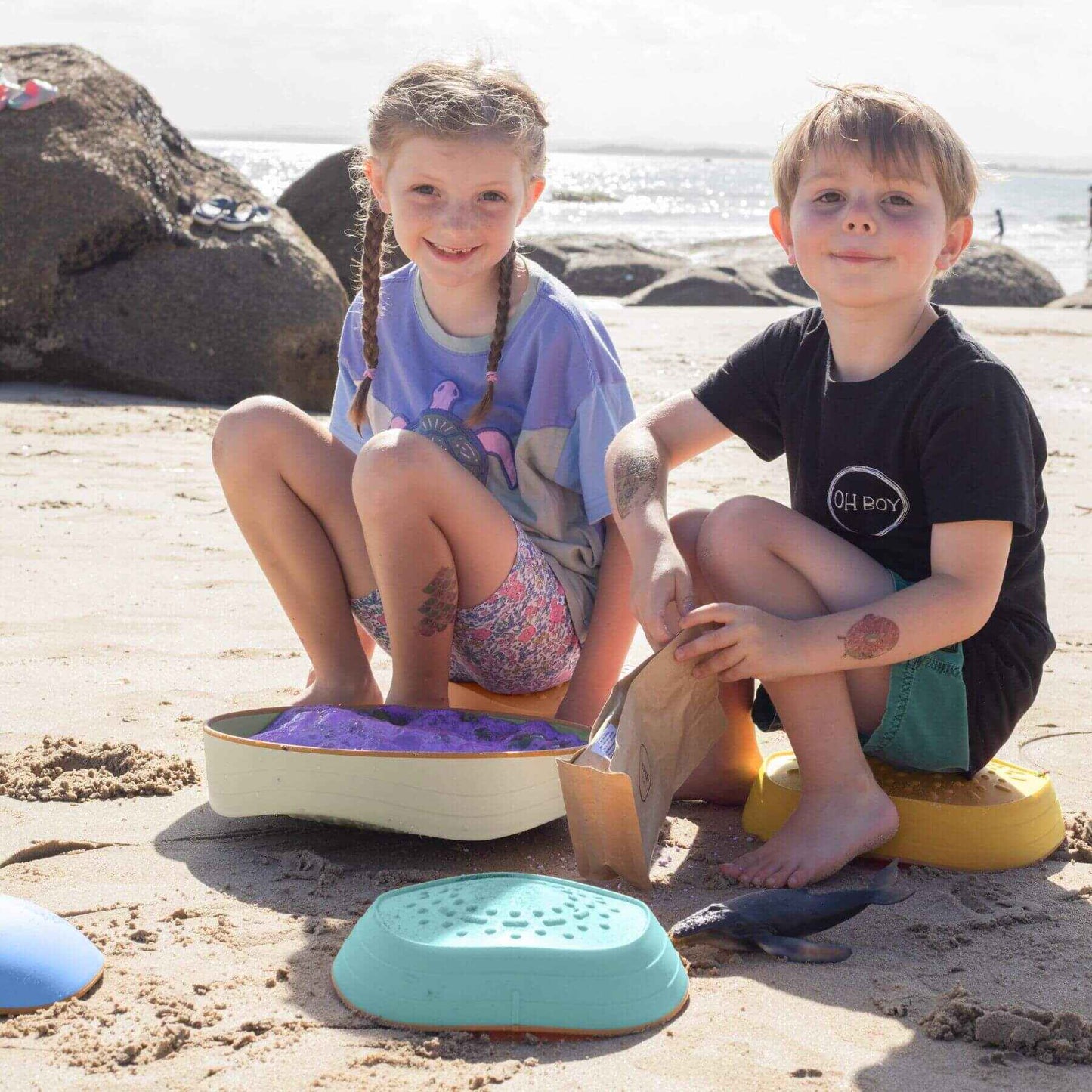 Two children sitting on Tiny Land® Stepping Stones - Beach Vacation in the sand with rocks and water in the background