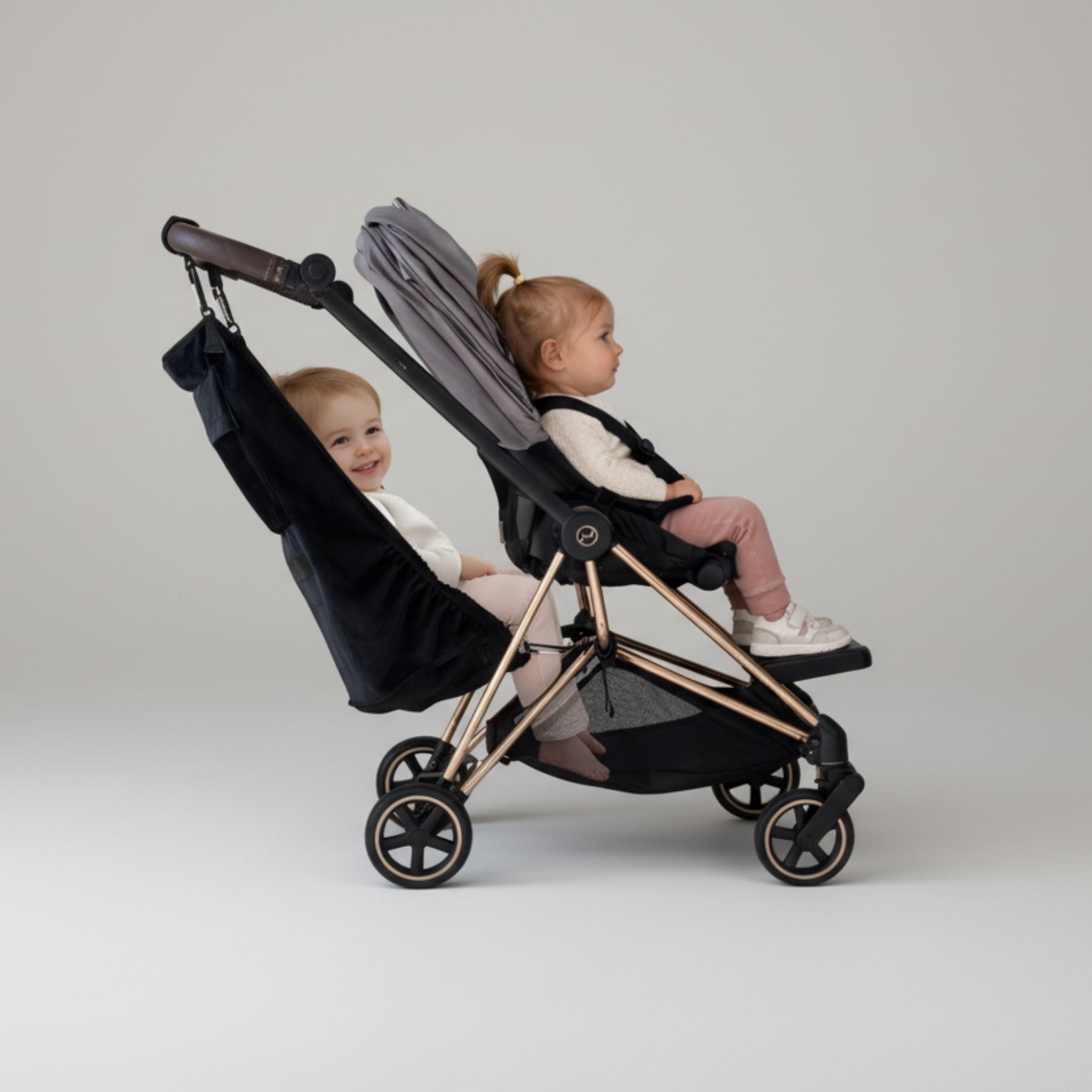 Two children sitting in a black and rose gold stroller against a plain background