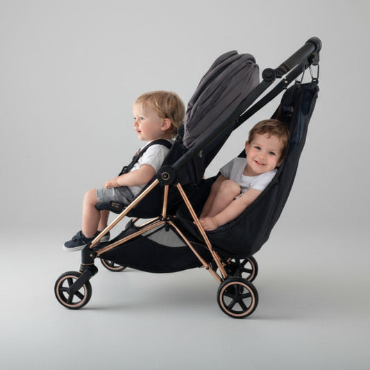 Two children sitting in a black stroller with Stroller Sling™ – Black Fox Edition attachd, against a gray background