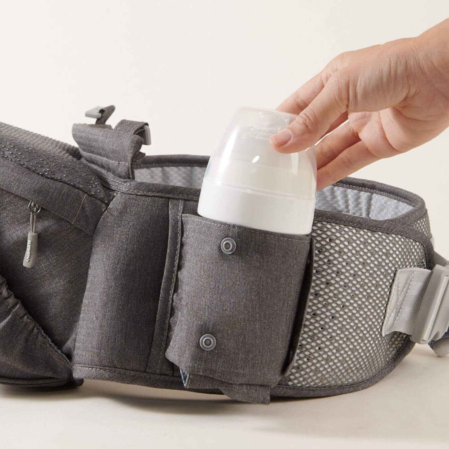 Sunveno Two Shoulder Straps Baby Hipseat with a hand placing a white bottle inside on a light background
