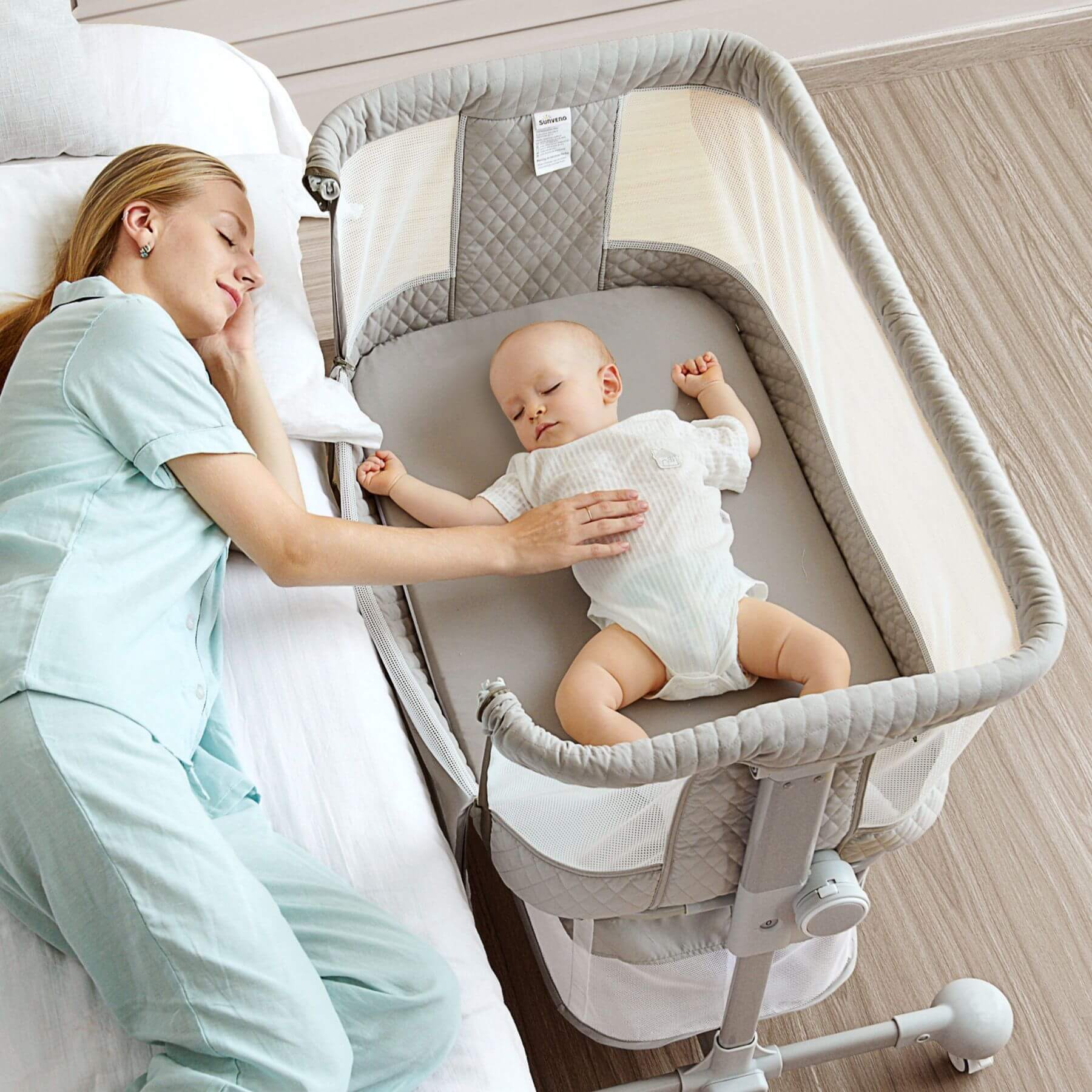 Woman holding a baby in Sunveno Baby Bassinet Bedside Crib next to a bed.