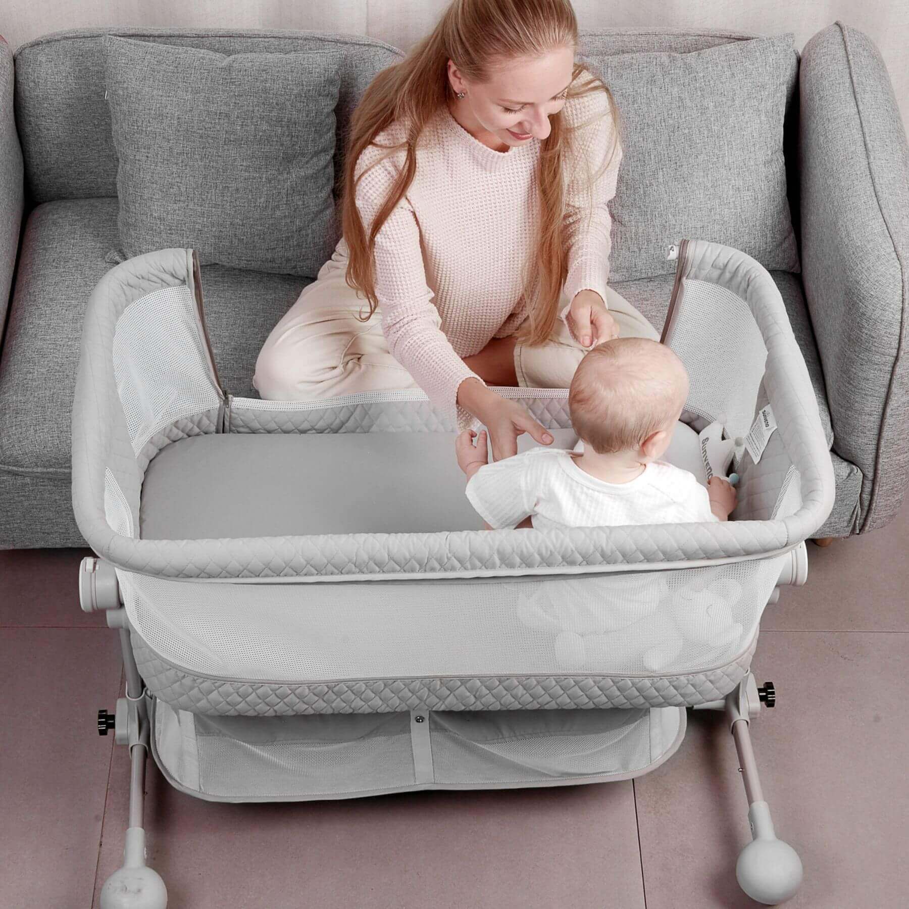 Woman sitting with a baby in Sunveno Baby Bassinet Bedside Crib in a living room.