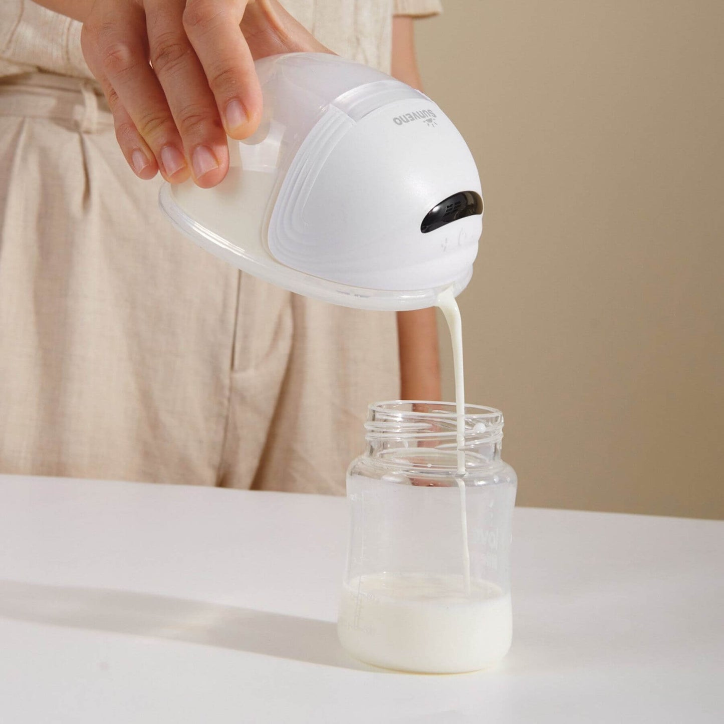 Person pouring milk from Sunveno Wearable Electric Breast Pump into a glass jar on a beige background