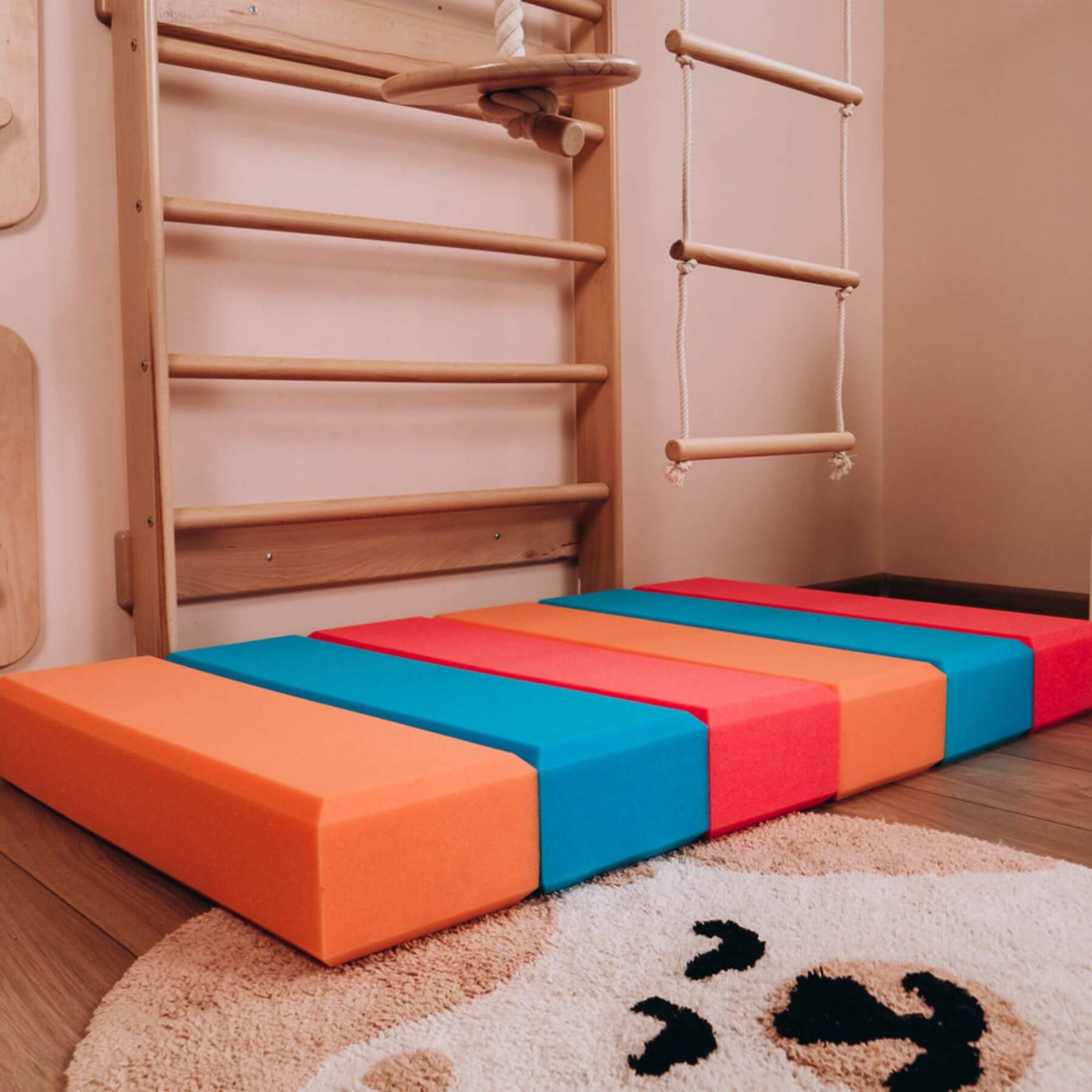 Children's play area with wooden climbing structure and colorful foam mats on a wooden floor.