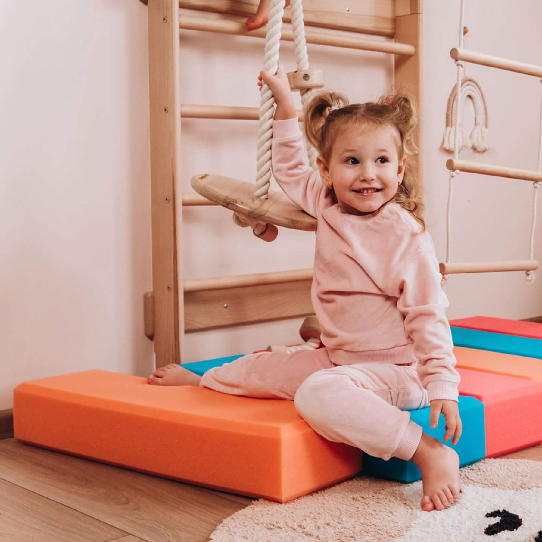 Child in pink outfit sitting on a colorful foam block in a playroom with climbing equipment.