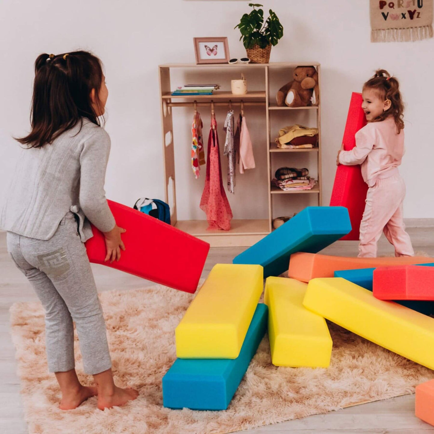 Two children playing with colorful foam blocks in a room with a shelf and clothes.