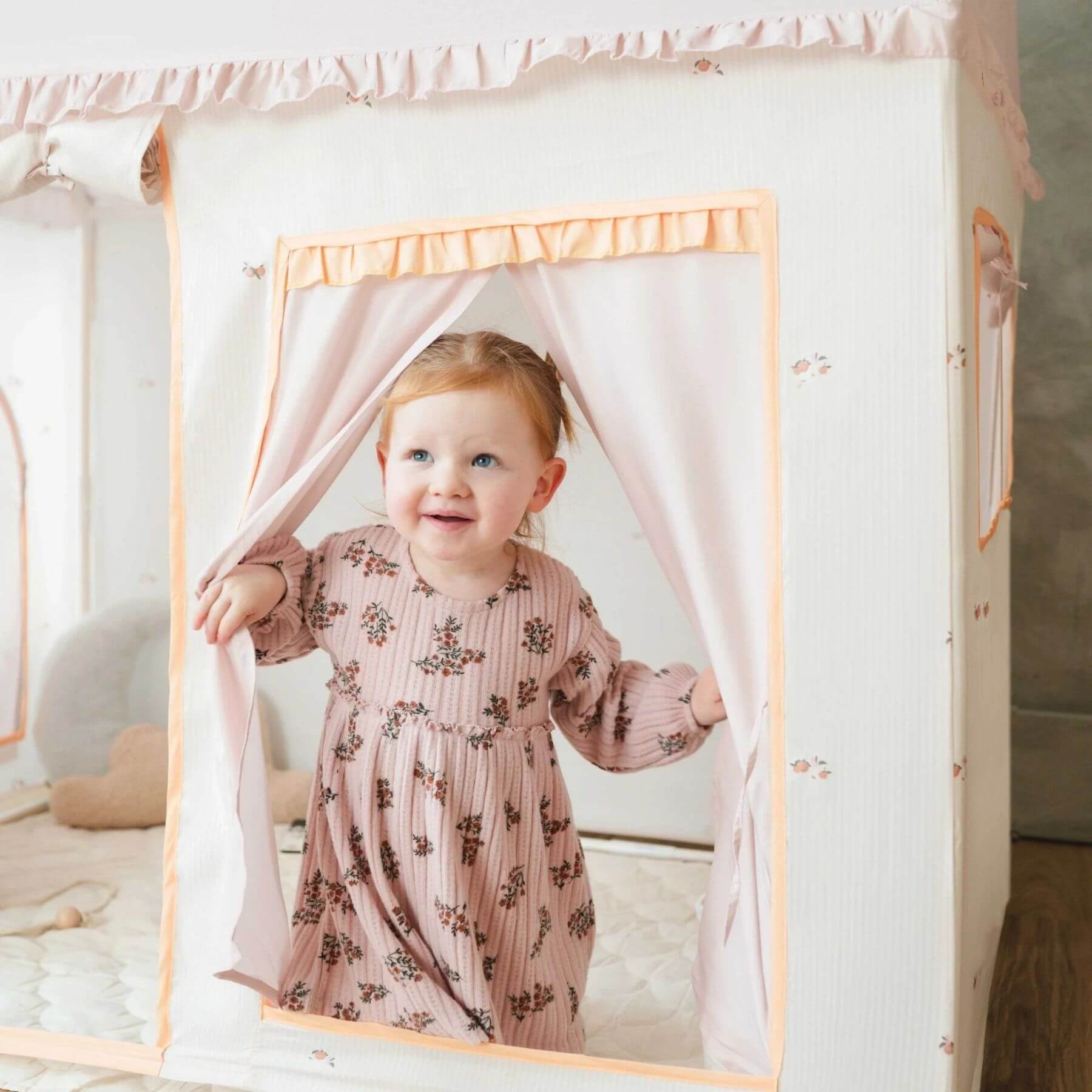 Child in a floral dress standing behind a white curtain with pink trim.