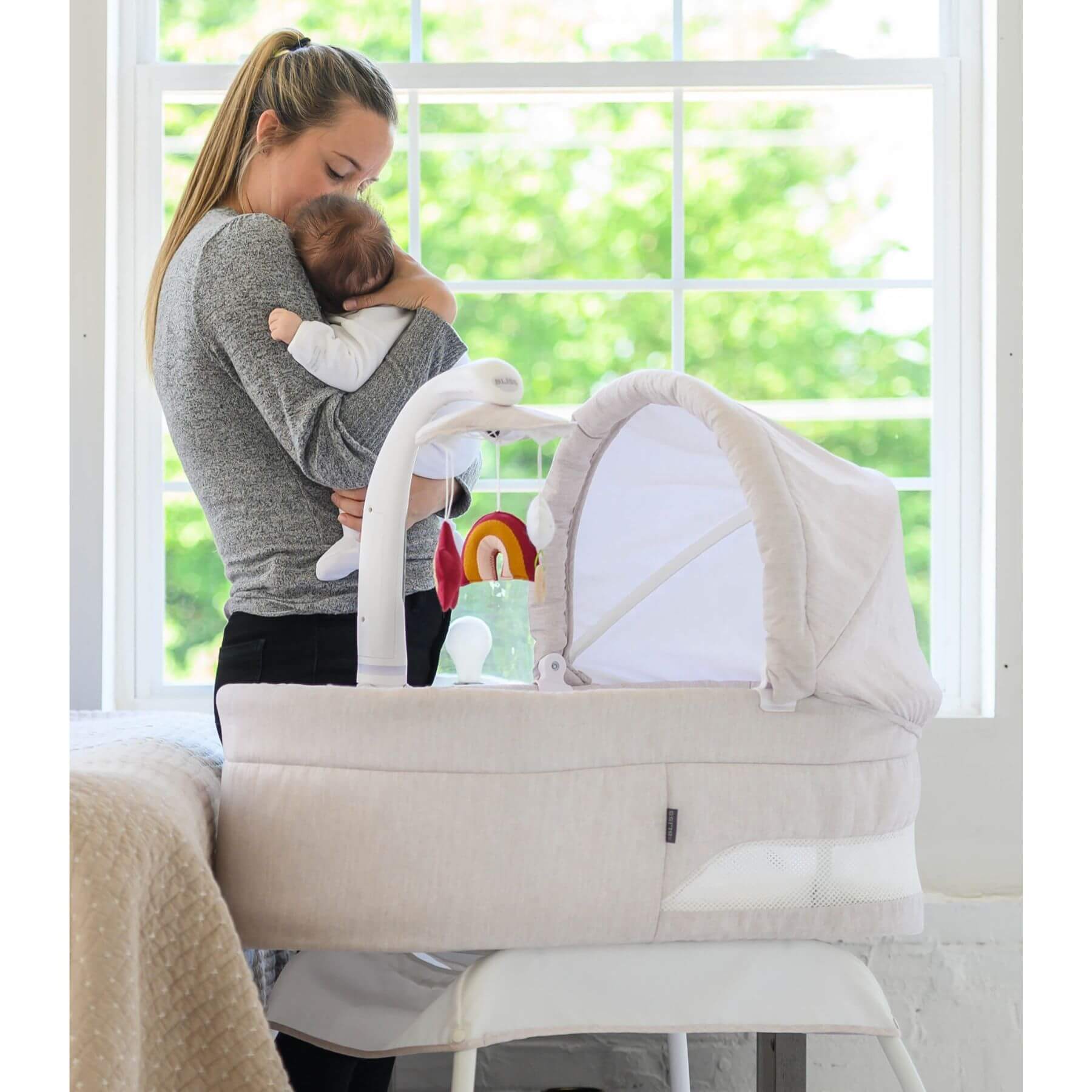 Mom Holding Baby next to TruBliss Sweetli Calm Bassinet Wheat Melange