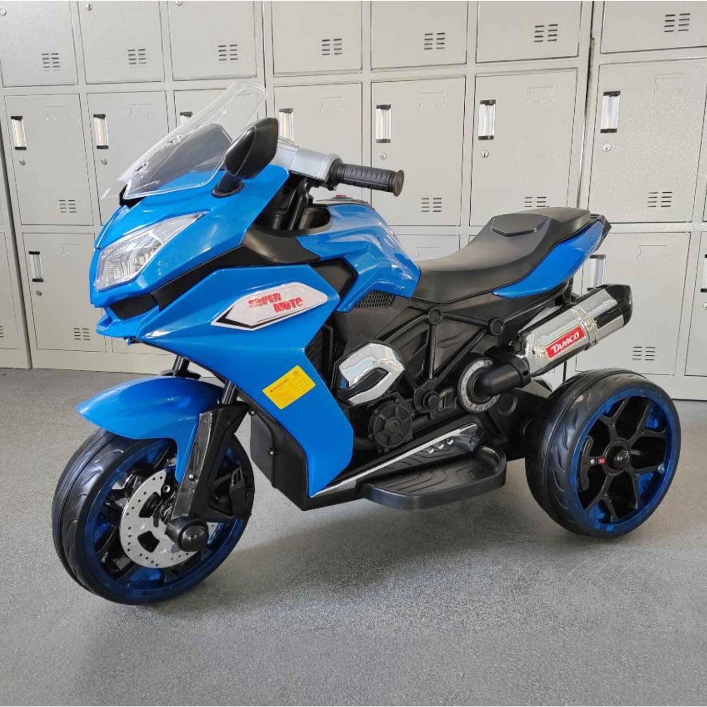 Tamco 12V Kids Electric Ride On Motorcycle - Blue on a gray floor in front of lockers.