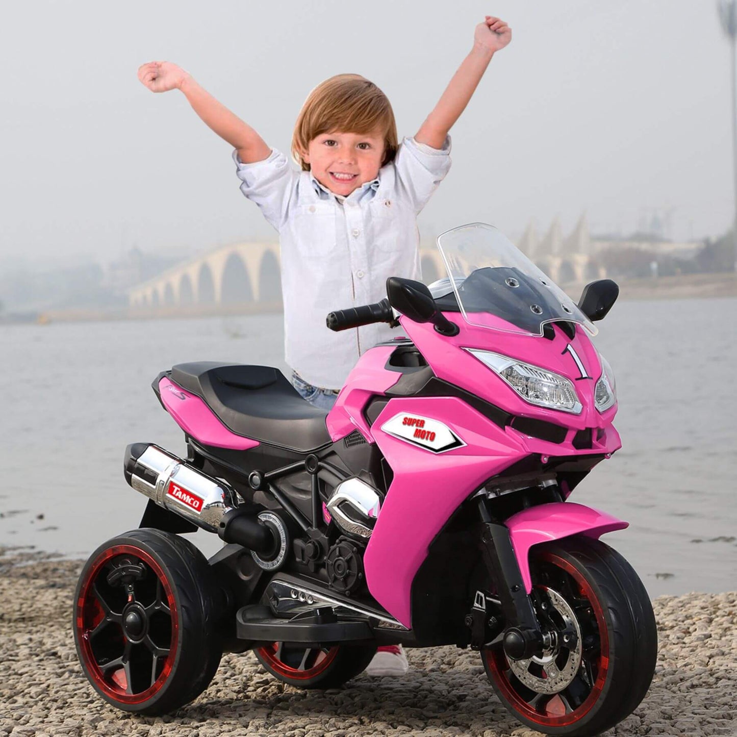 Child with arms raised next to Tamco 12V Kids Electric Ride On Motorcycle - Pink by a waterfront