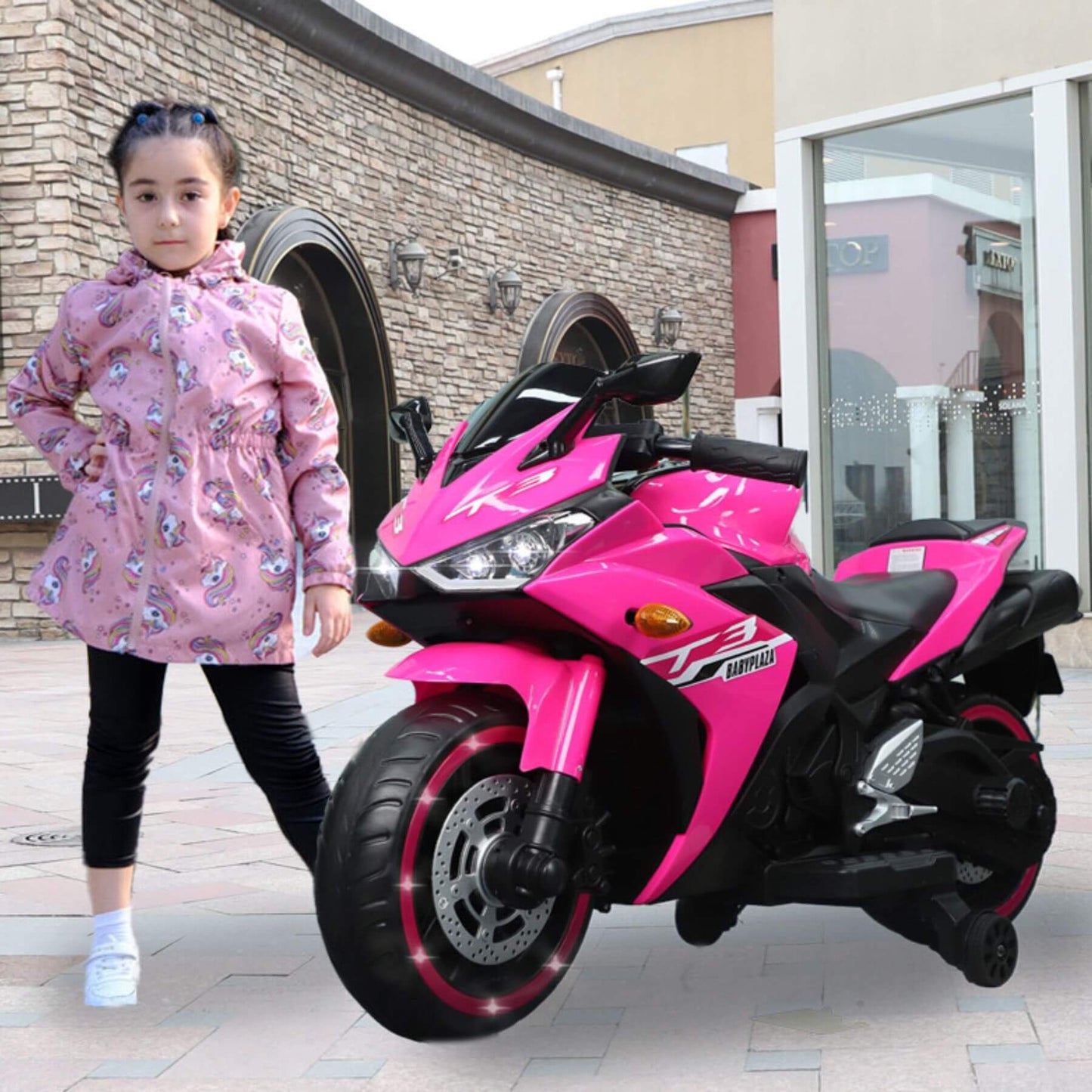 Child standing next to Tamco Kids 12V Motorcycle with Training Wheels - Pink on a patio.