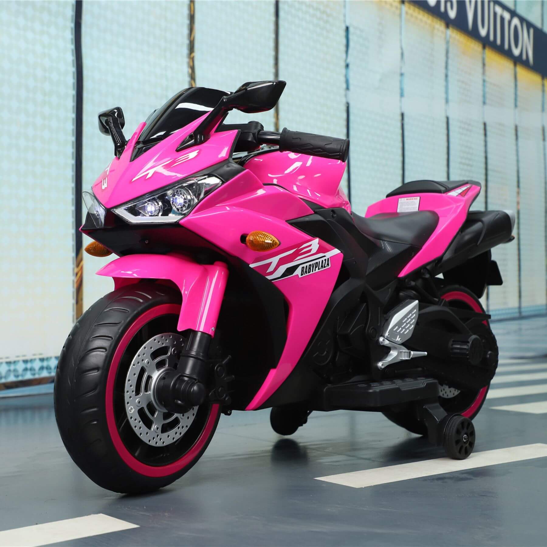Tamco Kids 12V Motorcycle with Training Wheels - Pink on a reflective floor with a blurred background