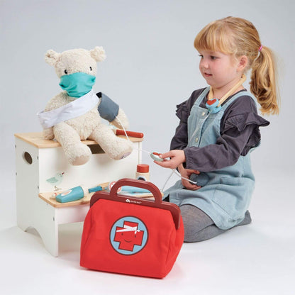 Child playing with a toy doctor set including a teddy bear, medical bag, and tools on a white background.