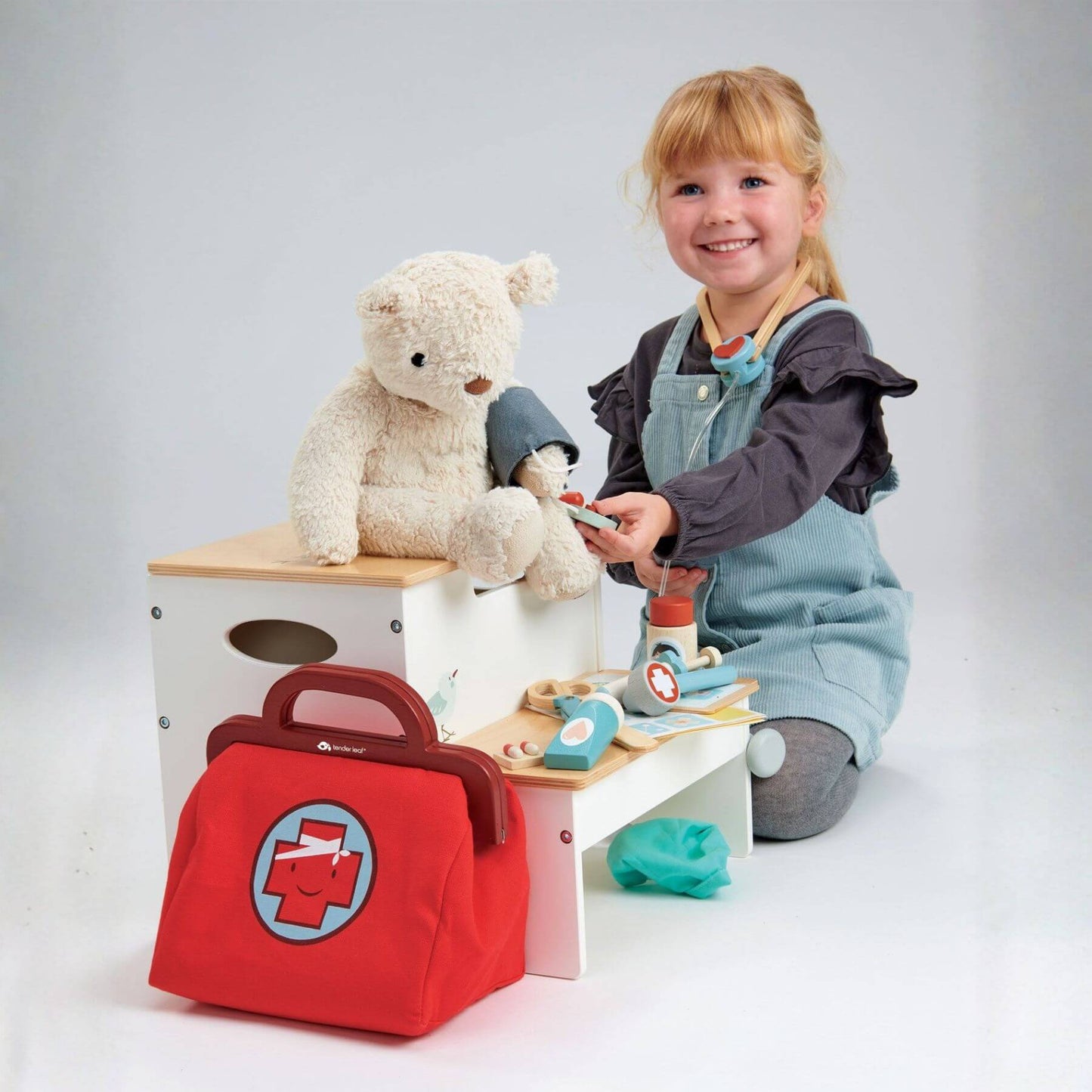 Child playing with toys including a teddy bear, toy medical bag, and wooden playset on a white background