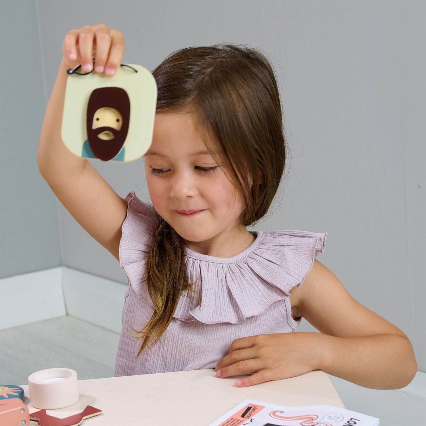 Young girl holding a toy with a face design, sitting at a table with a neutral background