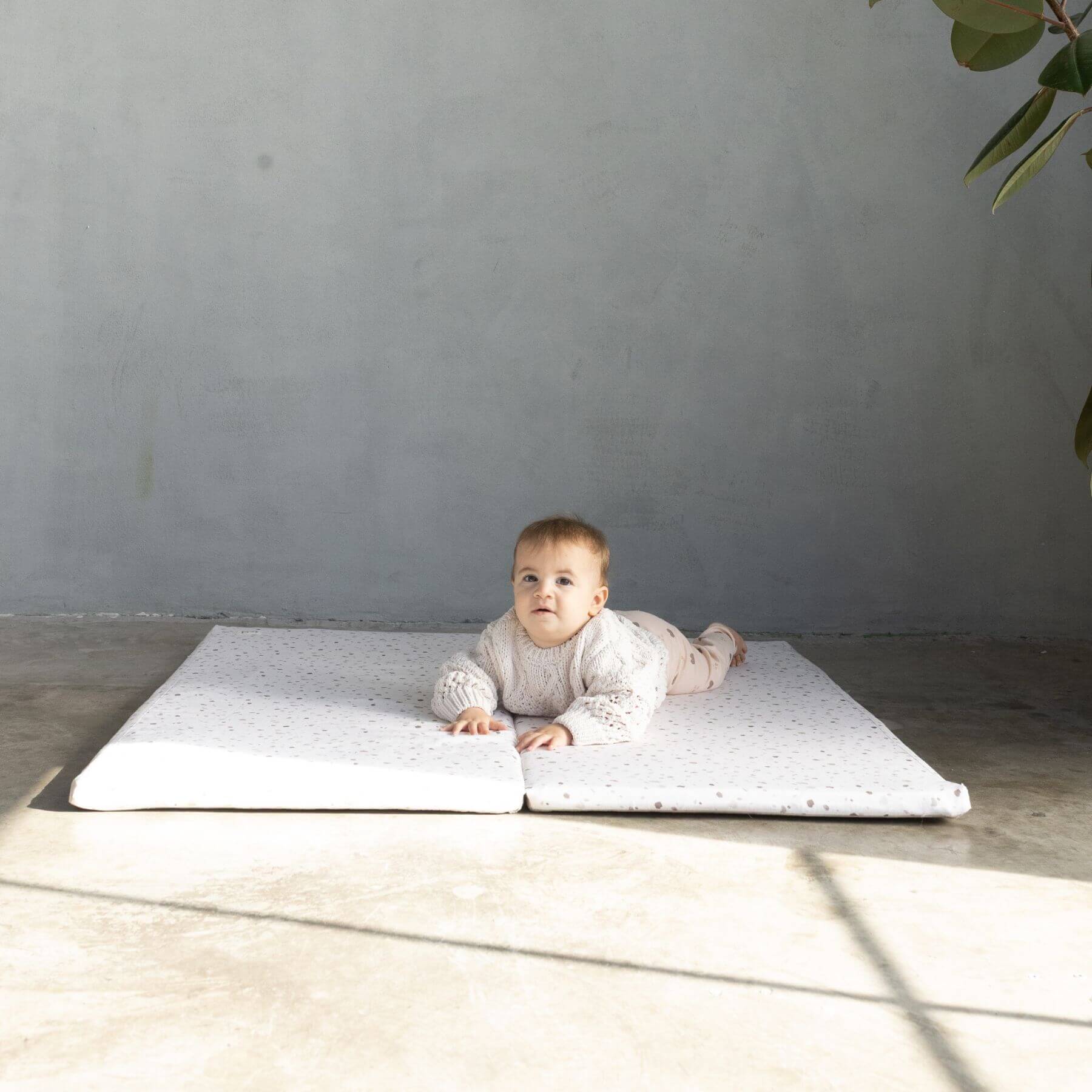 Baby lying on Terrazzo Chalk Organic Cotton Mat in a minimalistic room with a gray wall and wooden floor.