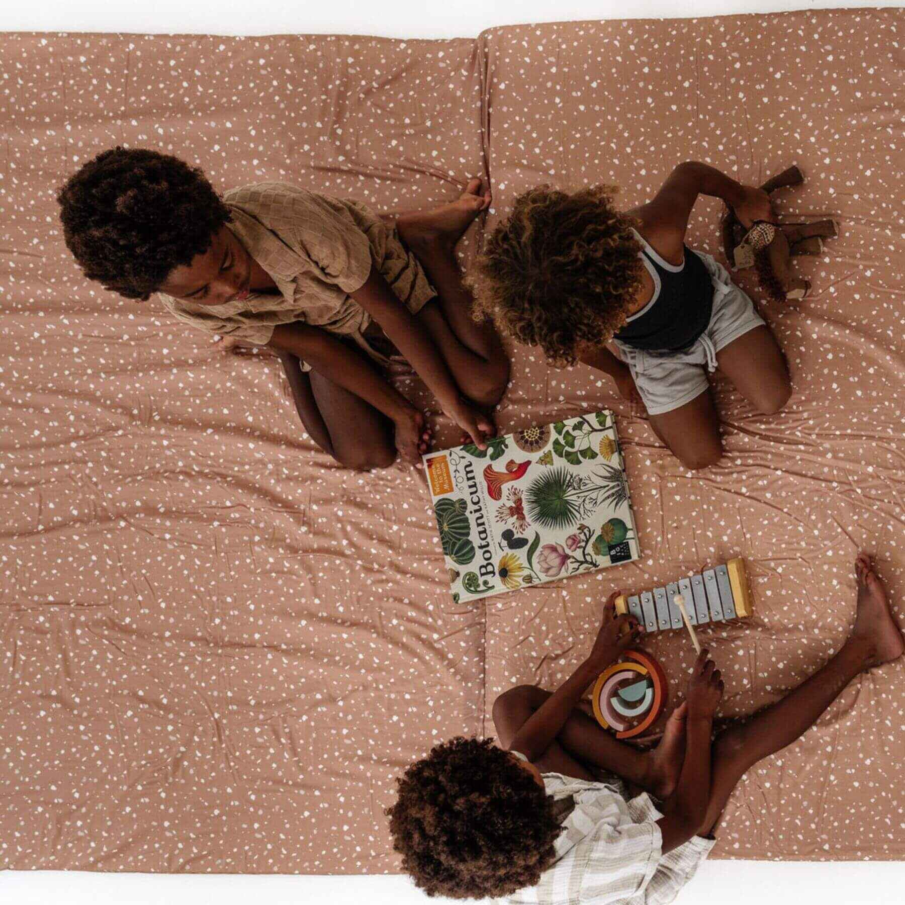 Three children sitting on Terrazzo Clay Organic Cotton Mat with books and toys around them.