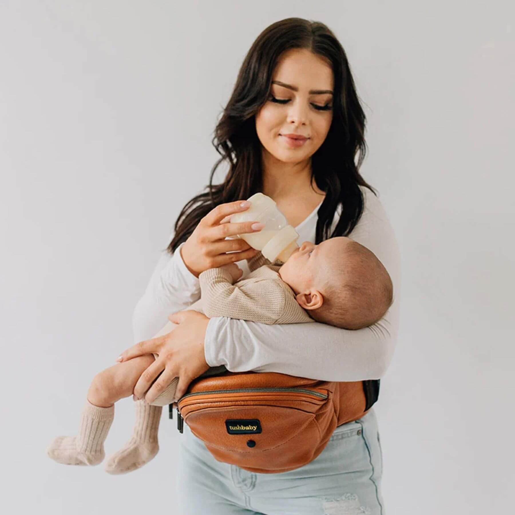 Woman holding a baby in The Tushbaby Hip Carrier - Vegan Leather Cognac