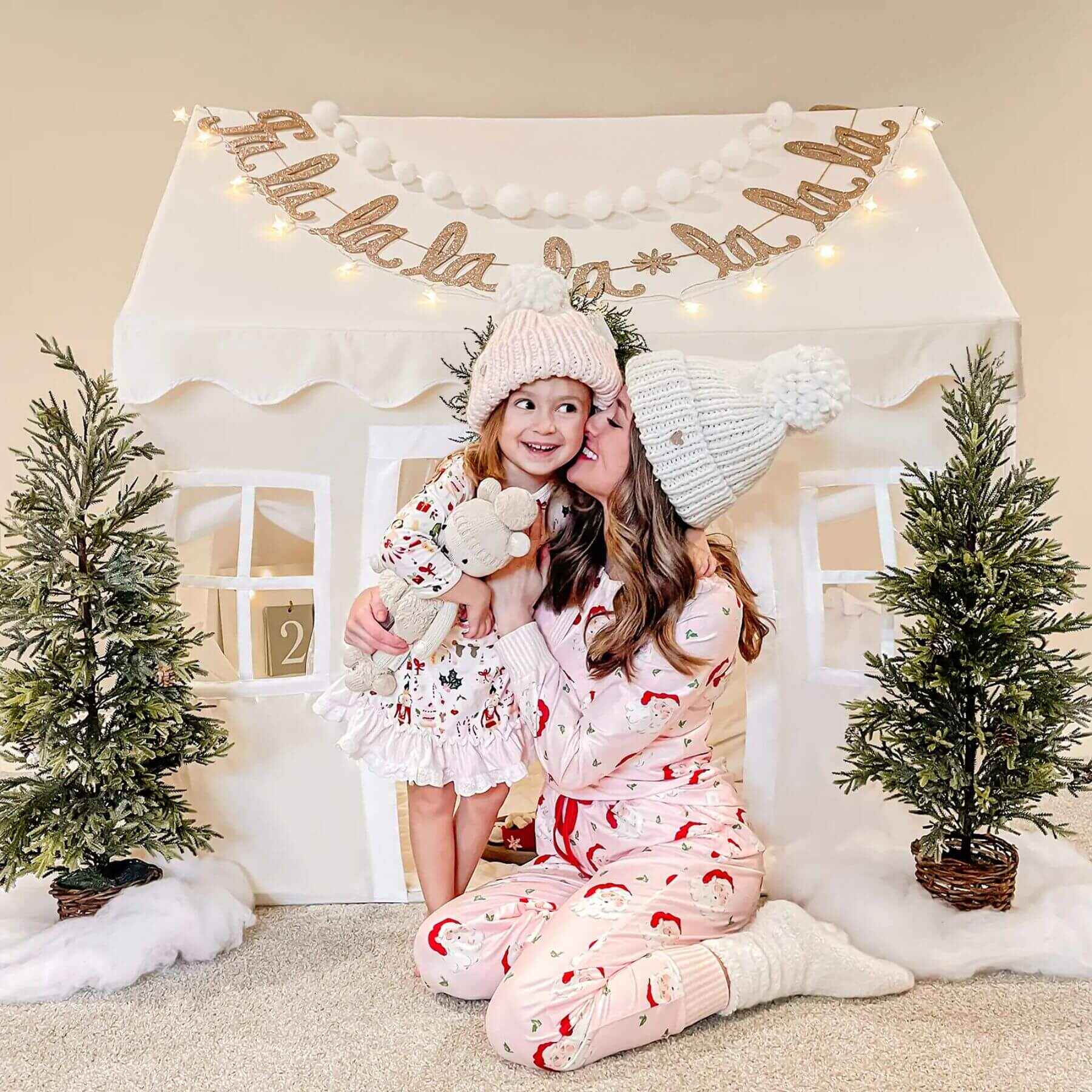 Two children in matching pajamas sitting in front of Tiny Land® Playhouse Tent – Natural Cream with Christmas trees and lights.