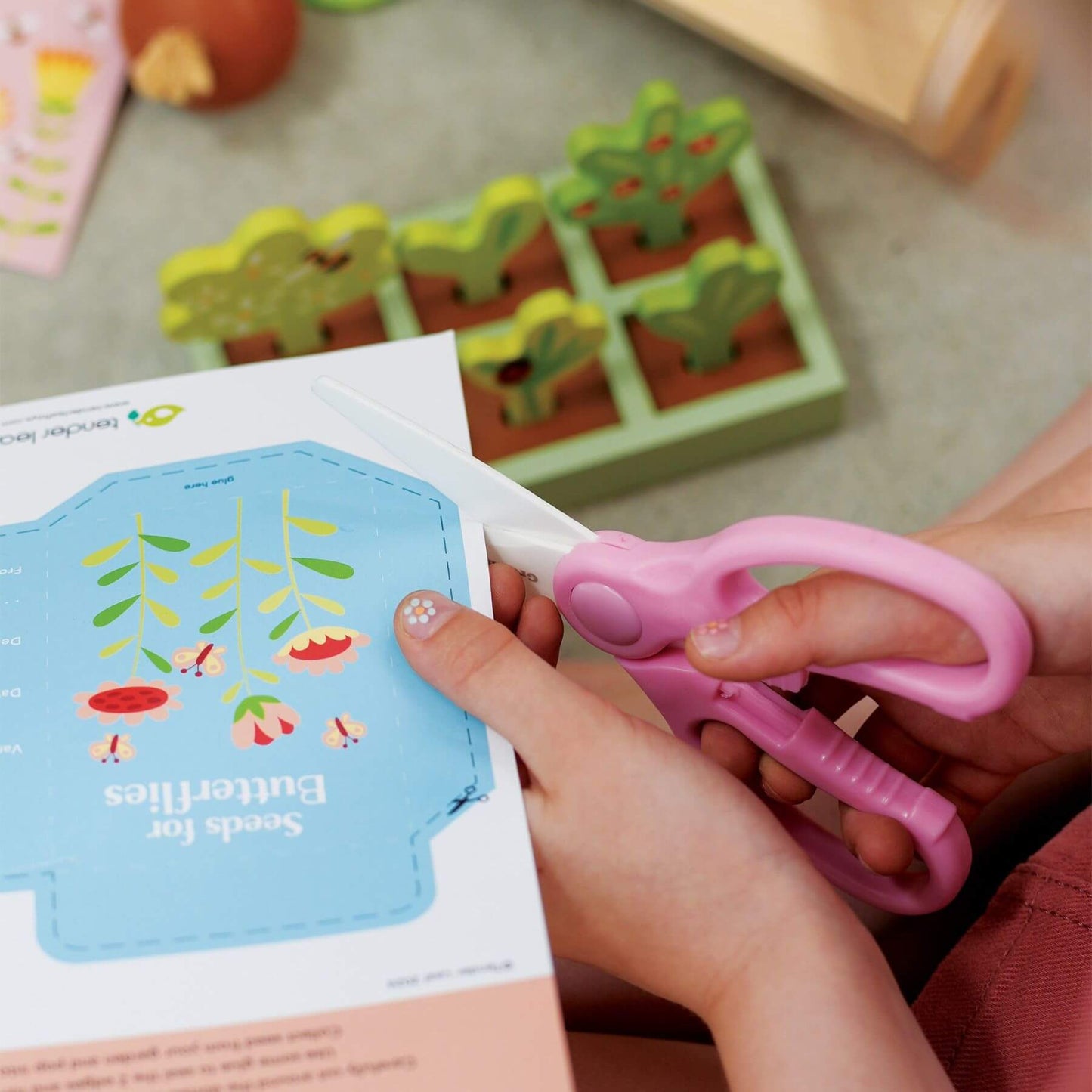 Child using pink scissors to cut out a butterfly craft from a book with a garden-themed puzzle in the background.