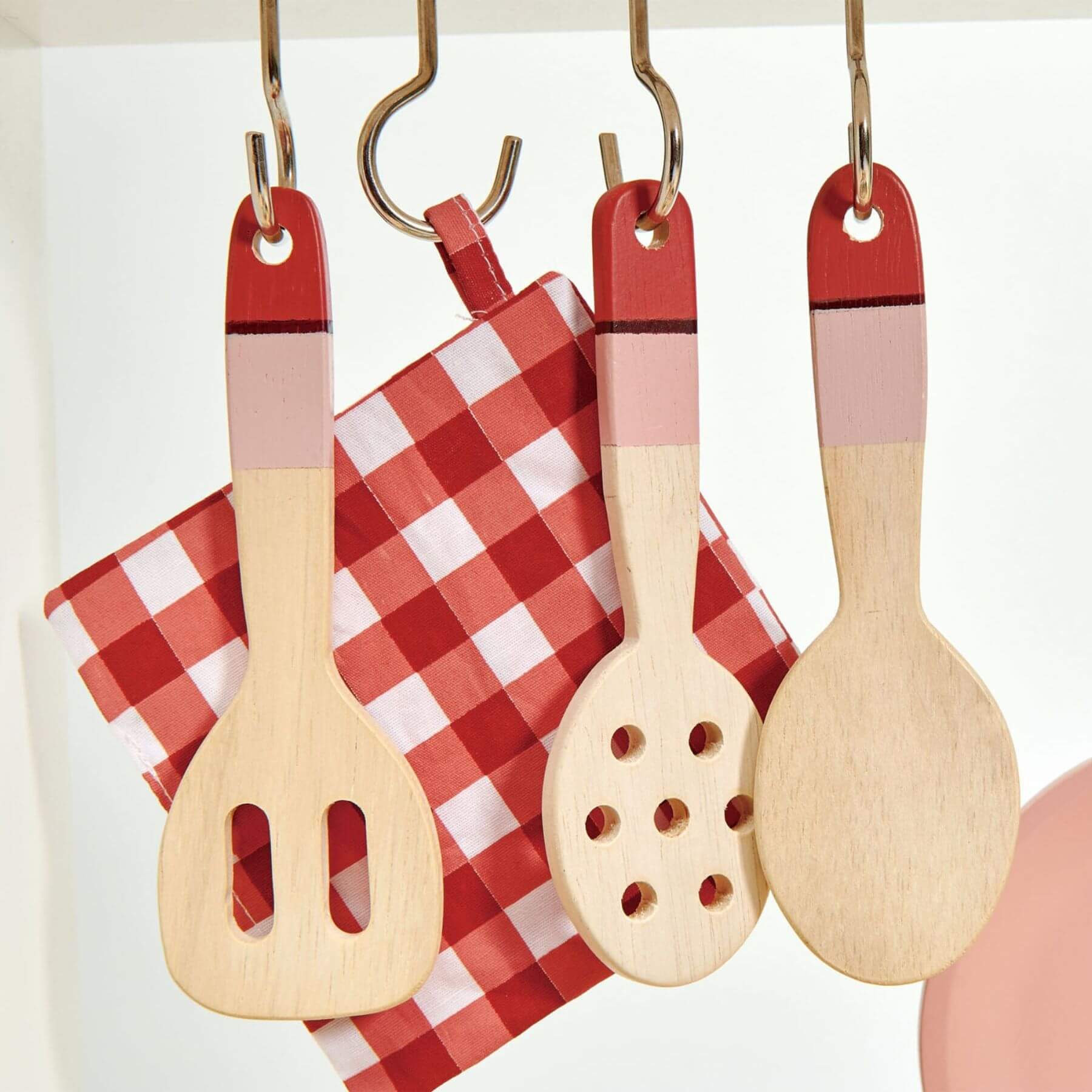 Set of toy wooden kitchen utensils with red and beige handles hanging on a hook against a white background.