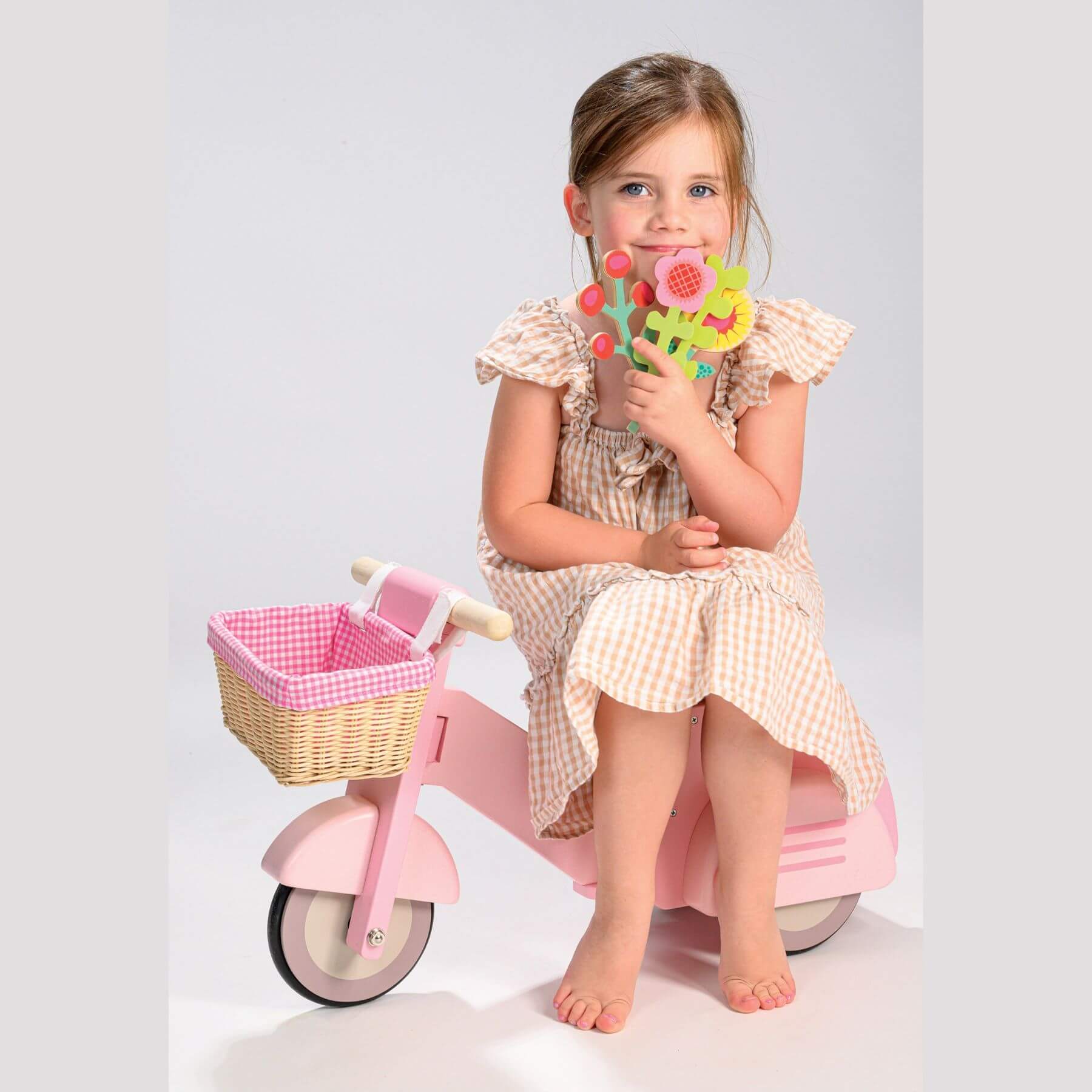 Young girl sitting on Tender Leaf Scooter with Basket, holding a toy, against a light gray background