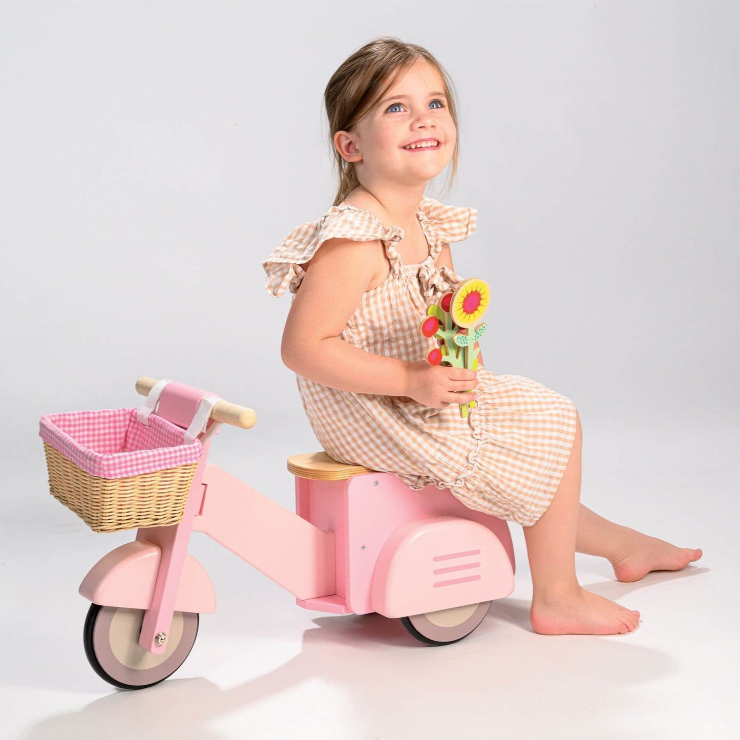 Young girl sitting on Tender Leaf Scooter with Basket holding flowers against a plain background