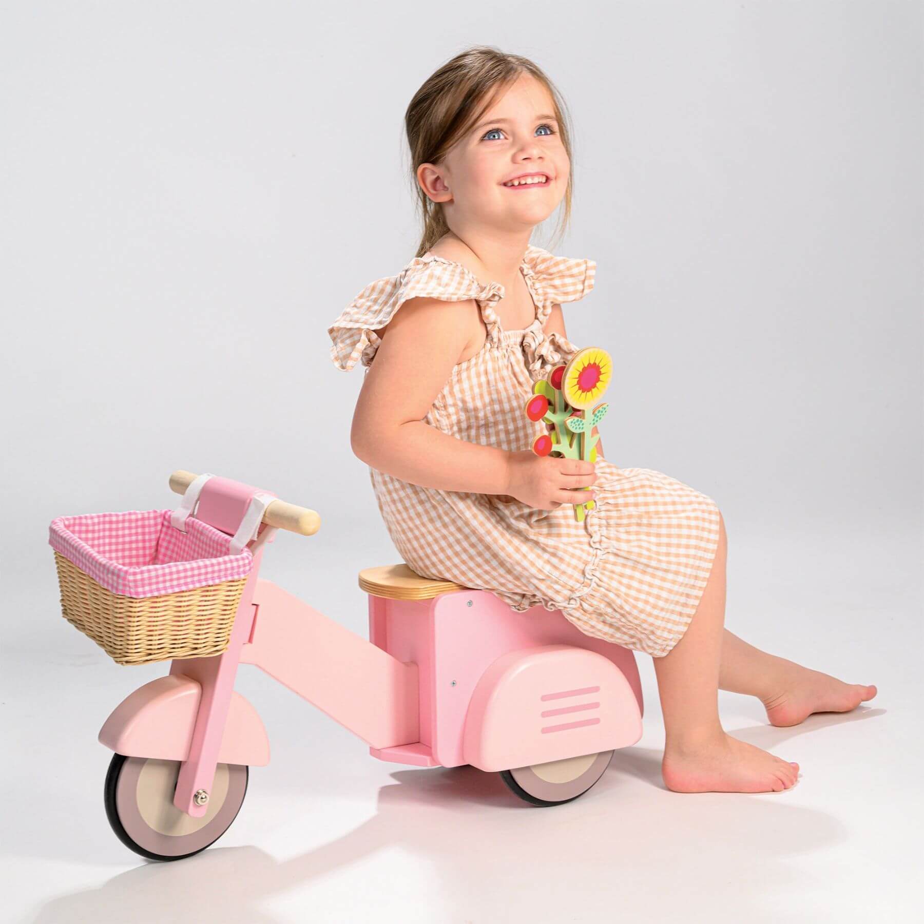 Young girl sitting on Tender Leaf Scooter with Basket holding flowers against a plain background